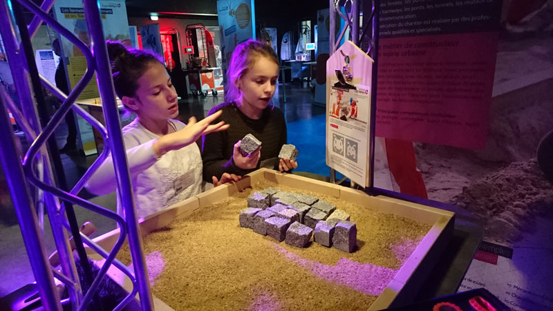 Deux jeunes filles participent à un atelier de pavage sur sable, manipulant des pavés dans un espace d’exposition dédié au BTP.